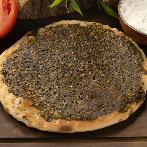 A round za'atar manakish on a wooden surface, with a tomato slice and flour bowl in the background.