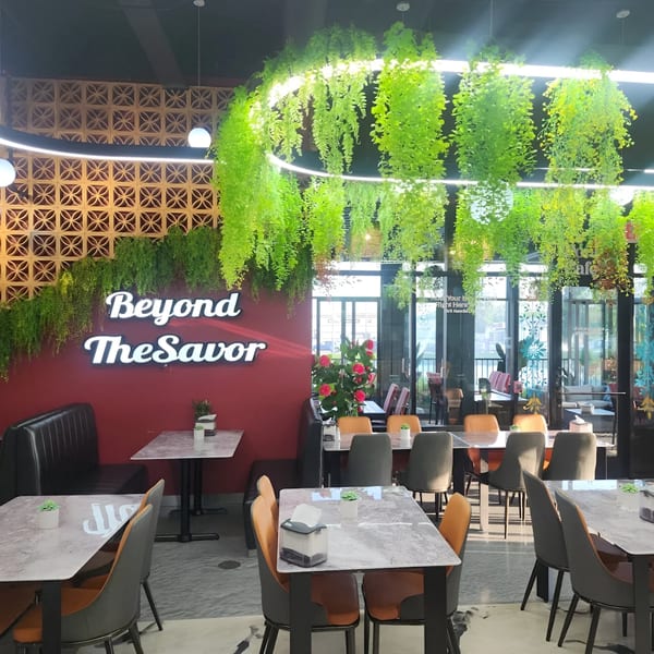 The Baba Saj dining room features modern tables and chairs, a red wall with "Beyond The Savor" neon sign, and hanging greenery.