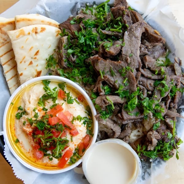 Baba Saj's Mediterranean dish with sliced meat, rice, hummus, pita bread, and a side of sauce.