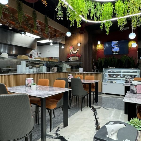 Interior of Baba Saj restaurant with dining tables, open kitchen, dessert display, and hanging greenery.