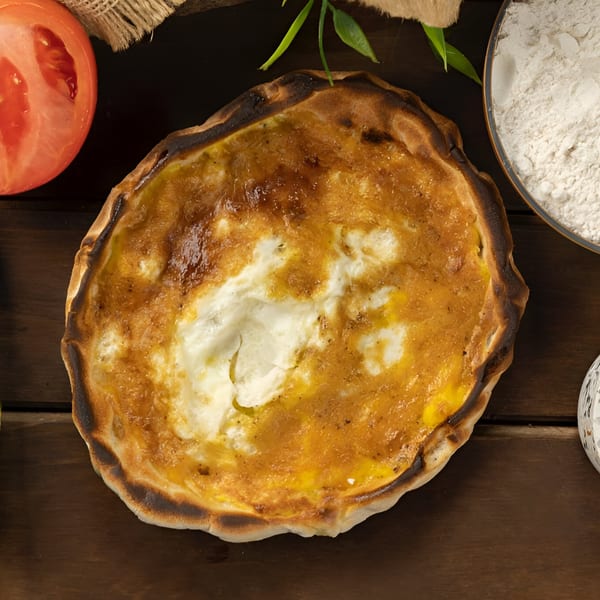 A golden-brown Mediterranean cheese pie with a charred crust, alongside a tomato slice and flour.